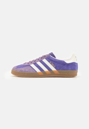 Fioletowy sneaker Adidas Gazelle z białymi paskami, gumową podeszwą i złotymi napisami z boku, pokazany z profilu na białym tle.