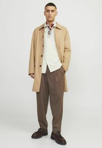 Beige frakke med sorte knapper, hvid skjorte, brune bukser og brune sko. Outfit inkluderer et rødt og grønt ternet tørklæde.