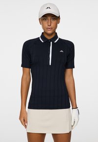 Polo da golf a maniche corte blu marina con texture rigata verticale, colletto e pattina bianca, logo ricamato, abbinato a una gonna beige e un guanto bianco.