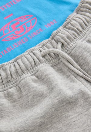 Sive trenirke z elastičnim pasom in vrvico za zategovanje, združene z modro majico z rožnatim napisom in okroglim grafičnim logotipom.