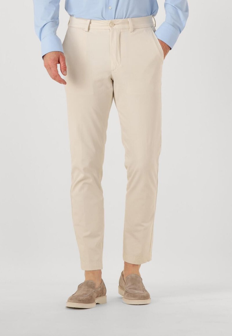 Man in beige slim-fit broek met zijzakken, lichtblauw shirt en taupe suède loafers, staande tegen een effen achtergrond.