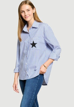 Jeune femme portant une chemise bleu et blanc rayée avec une étoile noire sur la poche poitrine et un jean bleu, souriant avec une main dans la poche.