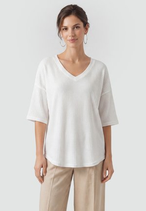 Mujer que lleva una camiseta blanca texturizada con escote en V y mangas hasta el codo, y pantalones beige, de pie frente a un fondo claro y liso.