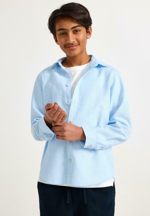 Teini-ikäinen poika hymyilee, hänellä on päällään vaaleansininen nappipaita valkoisen t-paidan päällä, ja hän säätää vasenta kalvostaan yksivärisen valkoisen taustan edessä.