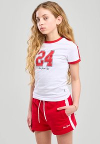Camiseta blanca con acentos rojos y gráfico "24" combinada con pantalones cortos rojos que tienen una franjas laterales blancas y cordón. Un atuendo casual y cómodo.