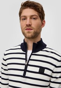 Striped cotton pullover in white and navy, featuring a zip neckline, ribbed collar, and a black logo patch on the chest.