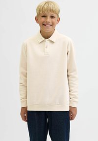 Beige långärmad polotröja med krage, tvåknapplåda och ribbade ärmslut, i kombination med mörka jeans. Slät textur.