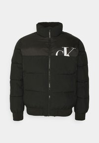 Sort dunjakke med høj krave, lynlås foran og elastiske manchetter. Har en kontrasterende blank panel og en stor hvid logo på brystet.