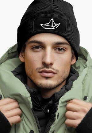 Black knit beanie with a white paper boat logo; model wears a green puffer jacket over a black inner layer, showcasing a casual look.