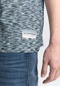 Šedá a černá košile s vzorem a štítkem s textem "TOM TAILOR", nošená přes modré džíny. Tkanina vypadá hladce s mírným leskem.