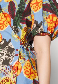 Robe en tissu floral multicolore avec manches bouffantes, présentant un fond bleu, des cercles jaunes, et une ceinture nouée à la taille. Texture lisse.