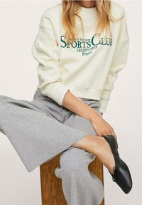 Felpa color crema con la scritta "SPORTS CLUB" in verde, abbinata a pantaloni della tuta grigio chiaro a gamba larga e ballerine nere. Seduta su uno sgabello di legno.