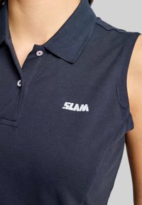 Marineblaues ärmelloses Poloshirt aus strukturiertem Stoff, mit einem weißen Logo auf der linken Brust und weißen Knöpfen mit roten Akzenten.