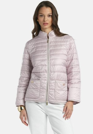 Žena v svetloružovej prešívanej bunde s prednými vreckami a zipsom, v kombinácii s bielymi nohavicami a zlatým náramkom.