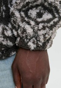Uldet sort og gråt sweater med hvide mønstre; tæt på tekstureret materiale, der viser det detaljerede design, med en brun hånd hvilende nedenunder.