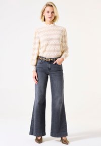 Spitzenbluse mit Puffärmeln und gewelltem Kragen in Creme, kombiniert mit hochgeschnittenen, ausgestellten Jeans aus dunklem Denim, akzentuiert durch einen Leopardenmuster-Gürtel.