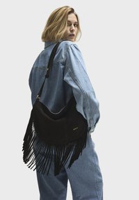 Black suede bag with long fringes, curved shape, and a gold logo. Worn over the shoulder with a strap. Model in denim outfit facing away.