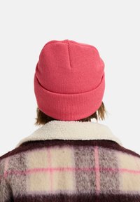 Roze gebreide beanie met een geribbelde textuur, gedragen door een persoon in een geruite wollen jas met een fleece kraag.