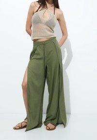 Pantalon ample léger vert olive avec fentes. Porté avec un haut halter en maille beige, présentant un design à trou clé. Sandales plates complètent le look.