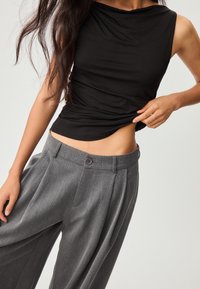 Schwarzes ärmelloses Oberteil mit einem drapierten Ausschnitt, kombiniert mit hoch sitzenden grauen plissierten Hosen, die einen vorderen Knopf und eine glatte Textur aufweisen.