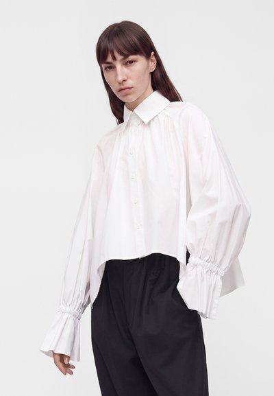 Femme aux cheveux bruns lisses portant une chemise blanche ample boutonnée avec des poignets froncés et un pantalon noir à jambes larges, sur un fond clair uni.