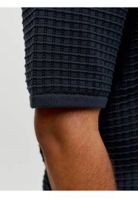 Gros plan d'un bras portant une chemise à manches courtes en maille bleu marine texturée avec un motif en grille et un poignet côtelé.