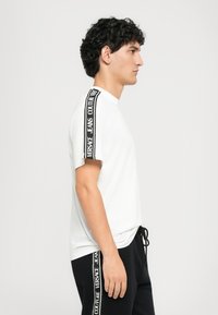 Jovem com cabelo encaracolado, vestindo uma camisa de manga curta branca e calças pretas com riscas do logotipo da Versace Jeans Couture nas mangas e laterais.