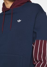 Mörkblå hoodie med maroonfärgad foderhuva, vit Adidas-logga och maroonärmar med vertikala vita ränder. Mjuk tygkvalitet, klassisk design.