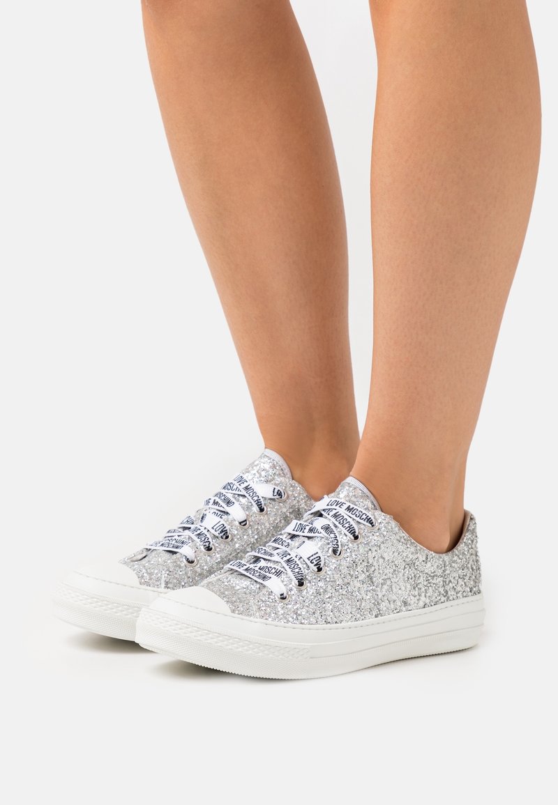 Pieds chaussés de baskets basses argentées pailletées avec semelles blanches et lacets blancs imprimés du texte « Love Moschino ».