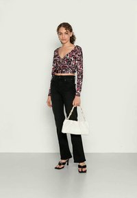 Blusa de manga longa em estilo envolvente com padrão abstrato em roxo e branco, combinada com jeans pretos de cintura alta, saltos pretos com tiras e uma carteira branca.