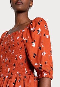 Vestido laranja rust com mangas bufantes, apresentando um corpete franzido e padrão floral em branco e preto. O tecido é leve e com uma textura suave.