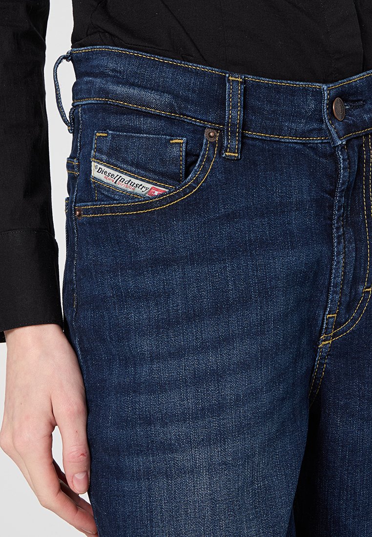 Jeans in denim blu scuro con cuciture gialle, una tasca frontale con un'etichetta e una vita a media altezza. La trama è liscia e resistente.
