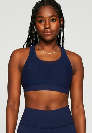 Frau mit geflochtenem Haar, die einen marineblauen Sport-BH und passende hoch taillierte Leggings vor einfachem Hintergrund trägt.