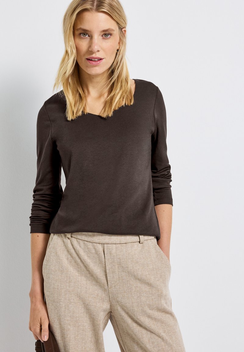 Ruskea pitkähihaisella paidalla, jossa on V-kaula-aukko, valmistettu sileästä kankaasta; yhdistettynä vaaleanruskeisiin teksturoituihin housuihin. Ei näkyviä kuvioita.