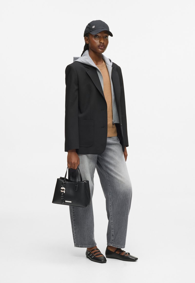 Γυναίκα φορώντας ένα μαύρο σακάκι πάνω από ένα γκρι φούτερ και μπεζ πουλόβερ, γκρι τζην, μαύρες μπαλαρίνες, μαύρο καπέλο, και κρατώντας μια μαύρη τσάντα.