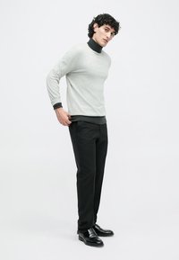 Szürke pulóver fekete garbóra rétegezve, fekete nadrággal és fényes fekete cipővel párosítva. Simább anyag, modern szabás, minimalista dizájn.