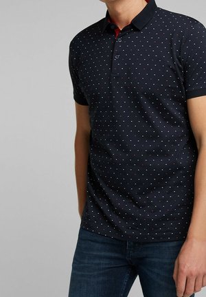 Mężczyzna w granatowej koszulce polo z małymi czerwono-białymi wzorami w trójkąty i ciemnoniebieskich dżinsach, stojący na szarym tle.