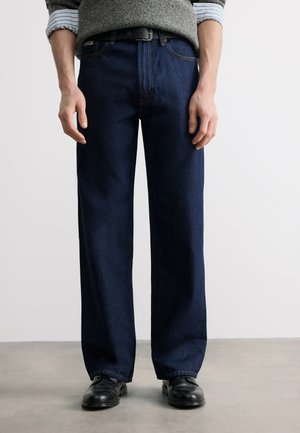 Uomo che indossa jeans a gamba larga blu scuro, cintura nera, maglione grigio sopra camicia a righe e scarpe di pelle nere, in piedi su pavimento grigio.