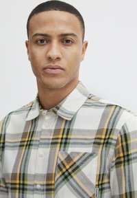 Camisa de botones con estampado de cuadros en colores blanco, verde, amarillo y gris; hecha de una tela ligera con un bolsillo en el pecho y collar.