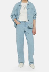 Světle modrá denimová bunda a odpovídající džíny, obojí se volným střihem. Bílé tričko s jemným grafickým logem pod ním. Bílé tenisky.