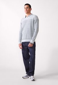 Helles graues Langarm-Polo mit schwarzen horizontalen Streifen, kombiniert mit dunkelblauen Jeans. Das Modell trägt weiße Sneakers und zeigt einen lässigen Stil.