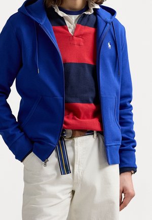 Persoon die een blauwe hoodie met rits draagt over een rood- en marineblauw gestreept rugbyshirt, witte broek met een gestreepte riem en een bruine leren riem.