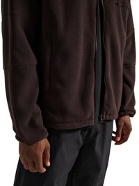 Homme portant une veste polaire marron foncé à fermeture éclair et un pantalon noir, montrant la texture de la veste et les poches avant.