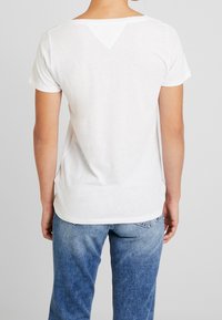 Camiseta blanca de manga corta con una espalda en pico, que presenta un discreto detalle de corte triangular en el escote, combinada con jeans de denim azul.
