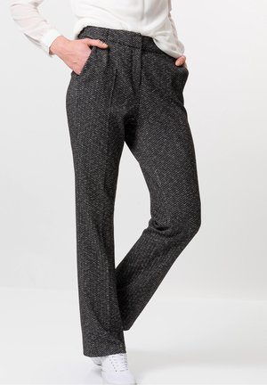 Person, die hoch taillierte, schwarz-weiße Hosen im Fischgrätmuster trägt, mit den Händen in den Taschen und weißen Turnschuhen, steht vor einem schlichten Hintergrund.