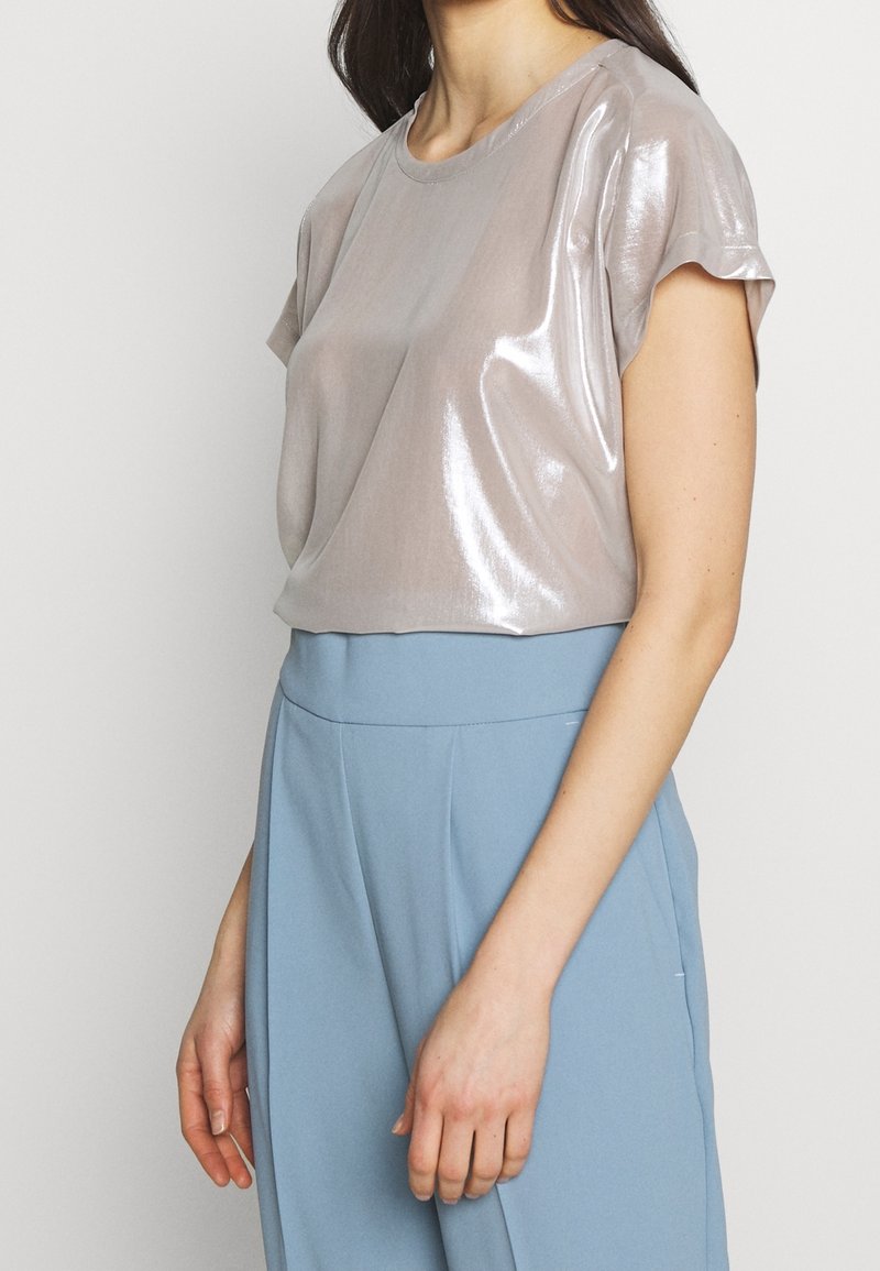Femme portant un haut court à manches courtes de couleur taupe clair brillant, rentré dans un pantalon large taille haute bleu ciel, sur un fond uni.