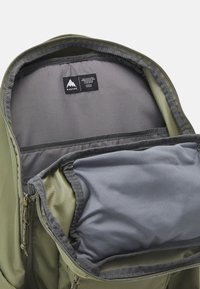 Intérieur d'un sac à dos vert avec doublure grise, comportant plusieurs compartiments et une étiquette de marque. Texture lisse, tissu durable et fermetures éclair présentes.