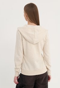 Felpa leggera beige con una consistenza morbida, cappuccio con cordino e polsini e orlo a coste. Indossata con pantaloni scuri, mostra un design casual.