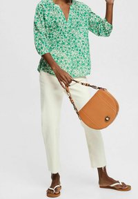 Blusa verde con motivo floreale e scollatura a V, pantaloni bianchi cropped con orlo sfrangiato e borsa in pelle marrone con tracolla a catena.