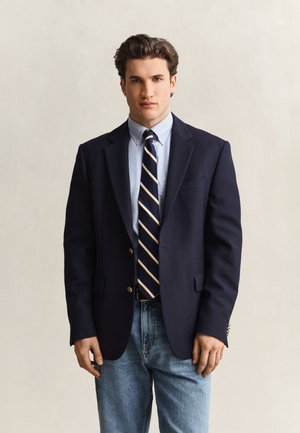 Uomo che indossa un blazer blu navy, camicia azzurra con bottoni, cravatta a righe e jeans blu, in piedi su uno sfondo chiaro e semplice.